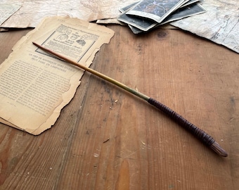 May include: A wooden wand with a brown leather wrap and a gold tip. The wand is lying on a wooden surface.
