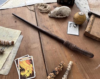 May include: A dark brown wooden wand with a carved handle lies on a wooden surface, alongside a skull, a yellow ball, and tarot cards. The wand is approximately 14 inches long. Other items include bundles of herbs and vintage ephemera.