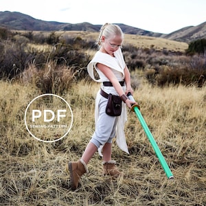 Könnte beinhalten: Ein junges Mädchen, das als Rey aus Star Wars verkleidet ist, trägt eine weiße Tunika und einen braunen Gürtel und hält ein grünes Lichtschwert in einem Feld aus trockenem Gras. Das Bild enthält den Text "PDF SEWING PATTERN".