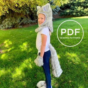 May include: A child wearing a grey wolf costume with a hood, tail and paw mitts. The costume is made of soft, fluffy fabric. The child is standing on a grassy lawn. The image includes the text "PDF SEWING PATTERN".
