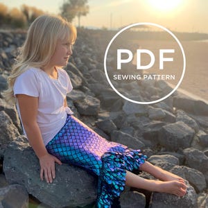 May include: A girl wearing a white shirt and a mermaid tail skirt with iridescent blue and purple scales. The skirt has a ruffle at the bottom. The girl is sitting on rocks by the water. The image has a circle with the text "PDF SEWING PATTERN" in white.