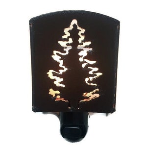 May include: A dark brown night light with a cut-out tree design. The tree silhouette is illuminated, creating a warm glow. The night light is rectangular with a rounded top and a black plug.