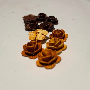 May include: A collection of small, wooden, flower-shaped embellishments in shades of brown, orange, and cream. The flowers are arranged in a semi-circle on a white surface.