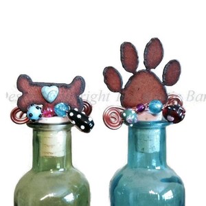 May include: Two decorative bottle stoppers. One features a brown bone shape with a blue heart, the other a brown paw print. Both have copper wire accents and colorful beads. The stoppers are on green and blue glass bottles.