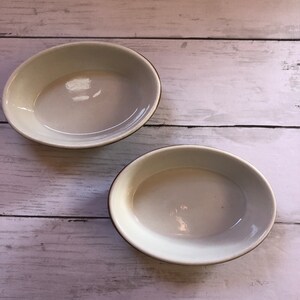 May include: Two white oval ceramic bowls with brown rims. The bowls are empty and sitting on a white wooden surface.