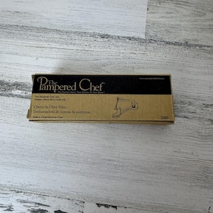 以下が含まれることがあります： キッチンツール用の長方形の段ボール箱、Pampered Chef Cherry & Olive Pitter。箱はタン色で、黒い文字とツールの線画が描かれています。テキストには会社の住所が含まれています。