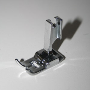 May include: A silver sewing machine foot with a curved needle plate and a raised metal shank.