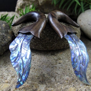 May include: Pair of dark wood ear gauges with iridescent blue abalone shell feather design. The gauges have a curved shape and are designed to be worn in stretched ear piercings. The abalone shell has a shimmering, multi-colored appearance.