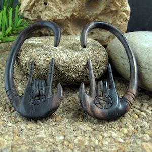 May include: A pair of black wood ear gauges with a carved hand design and a pentagram symbol. The gauges are shaped like a hoop and are made of dark wood.