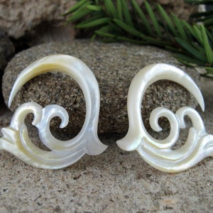 May include: A pair of white shell earrings with a carved, swirling design. The earrings are shaped like a crescent moon with a decorative flourish.