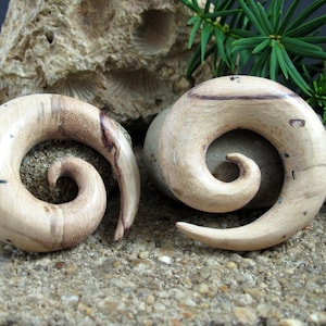May include: Two wooden spiral ear gauges, one with a light brown color and the other with a darker brown color. The gauges are carved with a spiral pattern and have a natural wood grain.