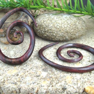 May include: Two dark red-brown wooden spiral earrings. The earrings have a smooth, polished finish and a swirling design. The earrings are displayed on a textured, light-coloured surface with a stone and greenery in the background.