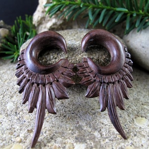 May include: A pair of brown wooden earrings with a carved feather design. The earrings are shaped like a crescent moon and have a long, pointed feather extending from the bottom.