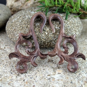 May include: A pair of brown wooden ear gauges with intricate, swirling designs. The gauges are shaped like a heart with a hook on each side.