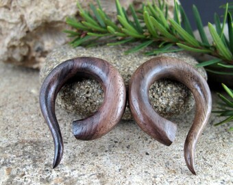 May include: A pair of dark brown wooden ear gauges, each shaped like a curved spiral. The gauges are made of a dark wood with a natural grain pattern.