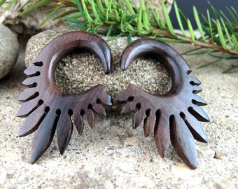 May include: Pair of dark brown wooden earrings with a feather-like design. The earrings have a curved, circular top and a series of carved, pointed shapes along the bottom edge. The wood grain is visible.