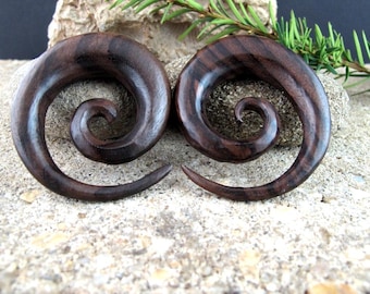 May include: A pair of dark brown wooden ear gauges shaped into smooth, polished spirals. Each piece features a tapered point that curves inward toward the center, showcasing the natural grain and deep, rich tones of the polished wood material.