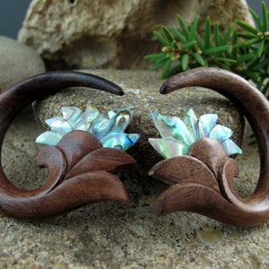 May include: A pair of brown wooden ear gauges with a carved floral design and iridescent abalone shell accents.