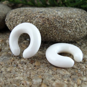 May include: Two white, smooth, rounded, horseshoe-shaped ear gauges. The gauges are made of a smooth, white material and are lying on a rough, gray surface.