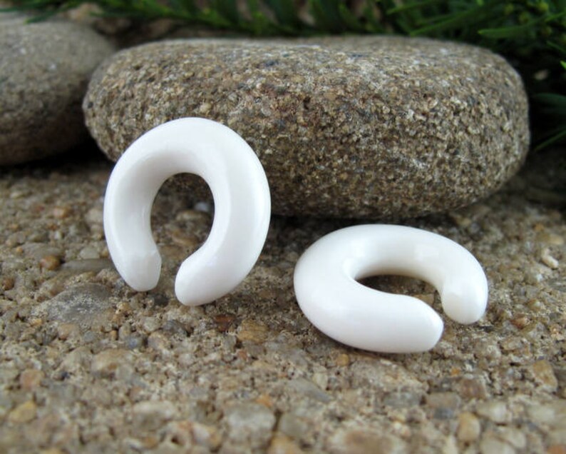 May include: Two white, smooth, rounded, horseshoe-shaped ear gauges. The gauges are made of a smooth, white material and are lying on a rough, gray surface.