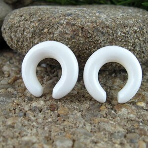 May include: Two white, smooth, horseshoe-shaped ear gauges. The gauges are made of a smooth, polished material and are a simple, elegant design.
