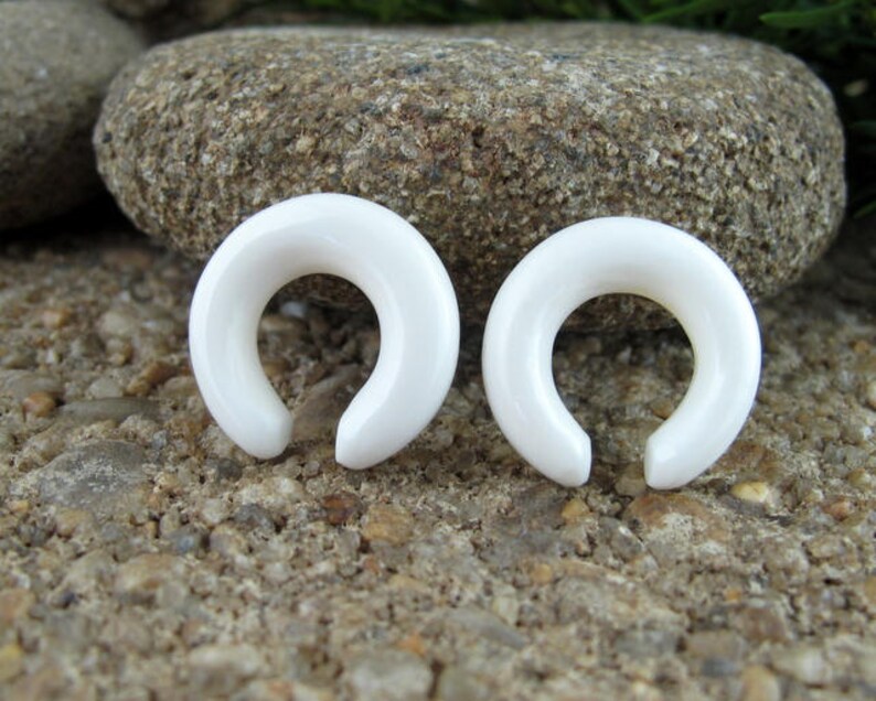 May include: Two white, smooth, horseshoe-shaped ear gauges. The gauges are made of a smooth, polished material and are a simple, elegant design.