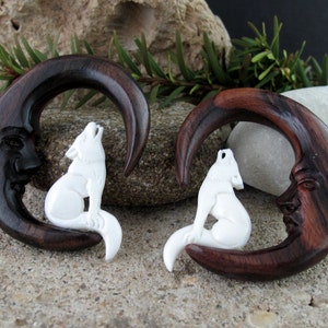 May include: Two dark brown wooden crescent moon plugs with carved faces, each featuring a white howling wolf. The plugs are designed for stretched ear piercings. The wood has a rich, dark grain.