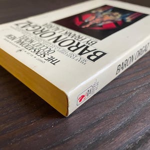 Pu&ograve; includere: Un libro bianco con una copertina dorata intitolato "Baron Orgaz" di Frank Lauria. La copertina del libro presenta un'illustrazione rossa e nera di una persona in un costume rosso e nero.