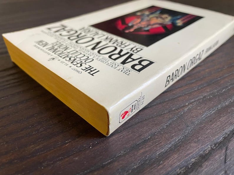 Pu&ograve; includere: Un libro bianco con una copertina dorata intitolato "Baron Orgaz" di Frank Lauria. La copertina del libro presenta un'illustrazione rossa e nera di una persona in un costume rosso e nero.