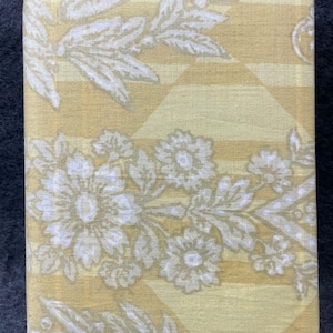 May include: A rectangular, yellow fabric-covered item with a floral and leaf pattern in white and light yellow. The design includes stripes and diamond shapes, creating a vintage aesthetic. The item is likely a book or a decorative box.