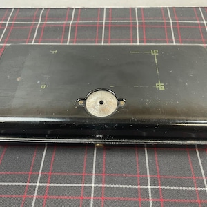 May include: A black metal toolbox with a circular, silver-colored knob on the top. The toolbox has a hinged lid and a small, rectangular handle.