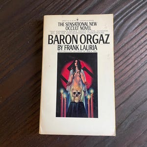 May include: A paperback book cover with a white background and black text. The title is "Baron Orgaz" by Frank Lauria. The cover features a woman in a white dress standing in front of a red swastika with candles and a skull.
