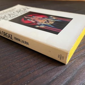 Pu&ograve; includere: Un libro tascabile bianco intitolato "The Confessions of Baron Orgaz" di Frank Lauria. La copertina presenta un'illustrazione in bianco e nero di una persona in costume rosso e nero.