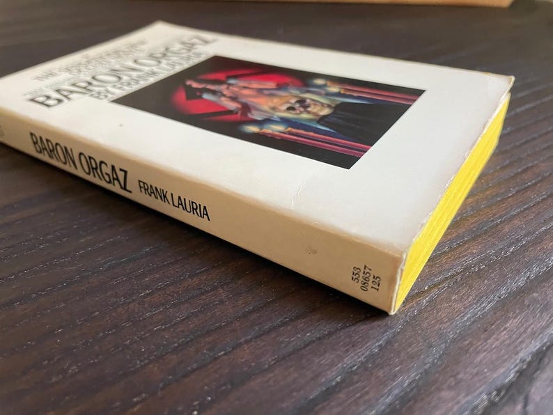 Pu&ograve; includere: Un libro tascabile bianco intitolato "The Confessions of Baron Orgaz" di Frank Lauria. La copertina presenta un'illustrazione in bianco e nero di una persona in costume rosso e nero.