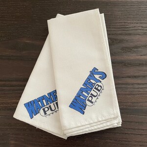 May include: Two folded white cloth napkins featuring the blue text "WATNEY'S PUB LONDON". The napkins are stacked on a dark brown wooden surface.