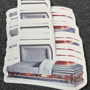 May include: A stack of stickers featuring a detailed illustration of a casket. The caskets are depicted in shades of brown, red, and blue, with a white interior and a white pillow. The stickers have a white border.