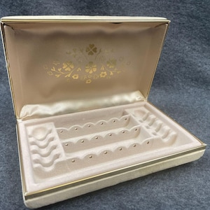 May include: Open jewelry box with a cream-colored exterior and a gold-colored trim. The interior is lined with a light beige fabric and features compartments for storing jewelry. The inside lid has a gold floral design.