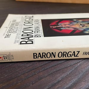 Pu&ograve; includere: Un libro tascabile bianco con il titolo "Baron Orgaz" di Frank Lauria. La copertina del libro presenta un'immagine in bianco e nero di una persona con un simbolo di svastica.
