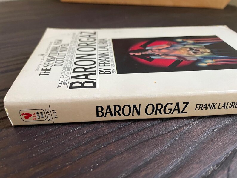 Pu&ograve; includere: Un libro tascabile bianco con il titolo "Baron Orgaz" di Frank Lauria. La copertina del libro presenta un'immagine in bianco e nero di una persona con un simbolo di svastica.