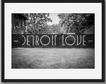 Puede incluir: Una impresión artística enmarcada en blanco y negro que muestra una valla de madera con el texto DETROIT LOVE en letras mayúsculas grandes. La escena incluye un patio con césped y árboles al fondo, presentada en un marco negro sobre pared gris.
