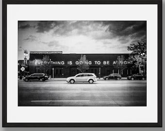 Puede incluir: Una fotografía enmarcada en blanco y negro que muestra el exterior de un edificio con el texto EVERYTHING IS GOING TO BE ALRIGHT en la fachada. Varios coches están aparcados frente a la estructura bajo un cielo nublado.