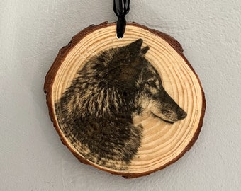 Puede incluir: Adorno redondo de madera con una imagen detallada de un lobo de perfil. El lobo está representado en tonos grises y marrones, sobre la veta natural de la madera. Se adjunta una cinta negra para colgarlo.