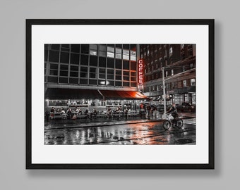 May include: A framed black and white photograph of a city street at night with red accents, featuring a coffee shop with outdoor seating, a cyclist, and wet pavement. The print is displayed above a black metal console table with books, a sphere, and an hourglass.
