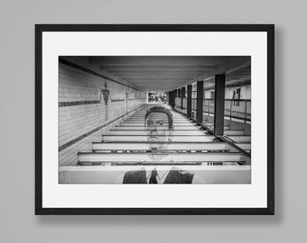 Puede incluir: Una fotografía en blanco y negro de un hombre de pie en un pasillo con paredes de azulejos y filas de bancos. El hombre lleva traje y corbata y mira directamente a la cámara. La fotografía está enmarcada en un marco negro y está colgada en una pared.