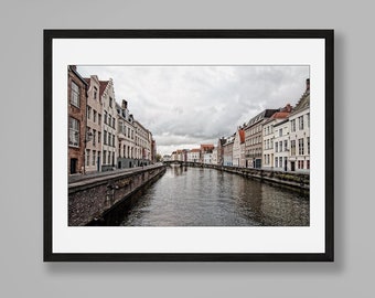 Puede incluir: Una fotografía de paisaje enmarcada de un canal bordeado por edificios históricos bajo un cielo nublado, sobre una mesa consola de metal negro con un reloj de arena de cristal con arena negra, un objeto esférico beige y libros blancos.