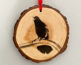 Puede incluir: Un adorno de madera con una silueta negra de un pájaro cantando en una rama. El adorno está sujeto a una cinta roja.