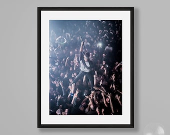 Puede incluir: Una fotografía en blanco y negro de una persona actuando en un escenario frente a una gran multitud. La persona está de pie sobre los hombros de la multitud, sosteniendo un micrófono en su mano. La multitud está levantando las manos hacia la persona.