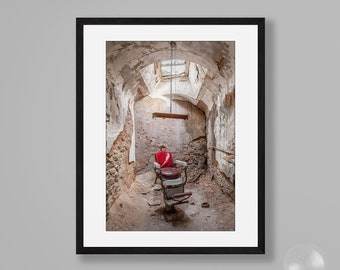 Puede incluir: Una silla de barbero roja se encuentra en una habitación deteriorada con pintura descascarada y una pared de ladrillo. La silla está suspendida del techo por una cadena. La habitación está iluminada por una pequeña ventana en el techo.