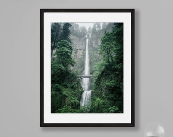 Puede incluir: Una fotografía enmarcada de una cascada que cae por un acantilado rocoso, rodeada de exuberantes árboles verdes. Un puente cruza la cascada, añadiendo un toque de presencia humana a la escena natural.