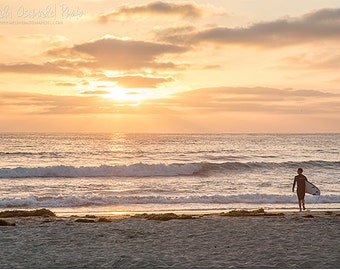 May include: A lone surfer walks along a sandy beach with a surfboard under their arm, as the sun sets over the ocean. The sky is a vibrant orange and pink, with fluffy clouds.